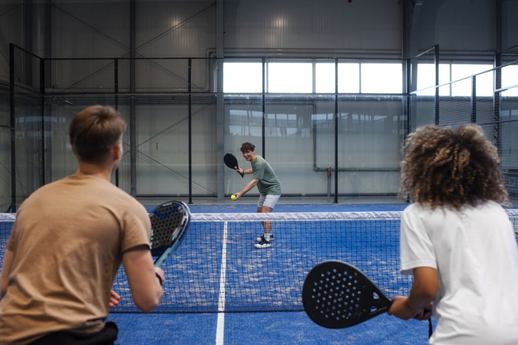 Sekelompok orang sedang tertawa sambil memegang raket padel di lapangan.